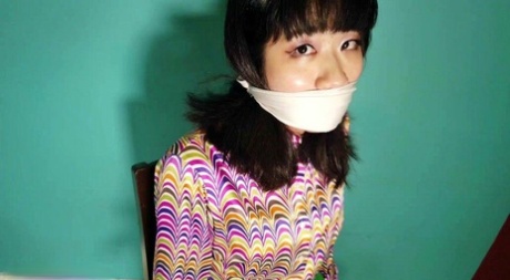 Oriental female applies a gag before handcuffing herself to a wooden chair 95440869